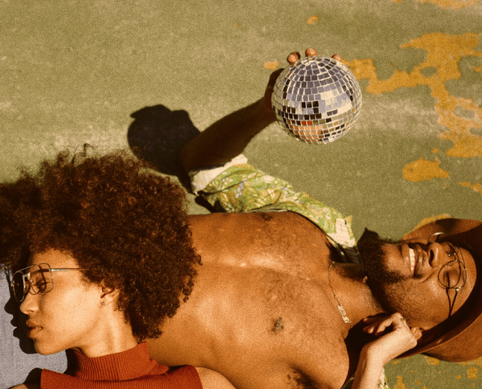 A couple lies on the ground, surrounded by the sparkling light of a disco ball, evoking a vibrant seventies atmosphere.